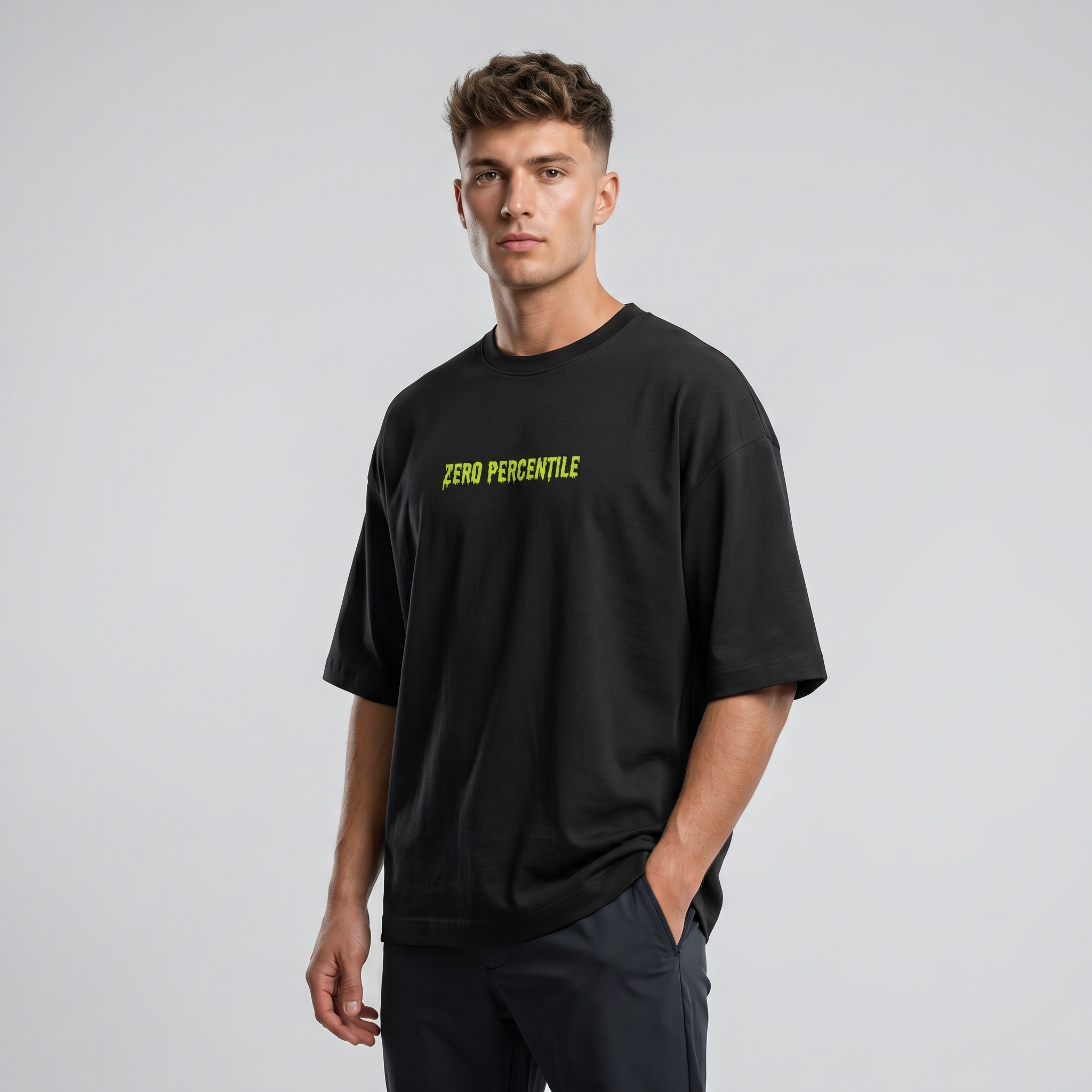 A man wearing a black oversized t-shirt with 'ZERO PERCENTILE' printed in neon yellow across the chest, paired with dark gray pants, against a light gray studio background.