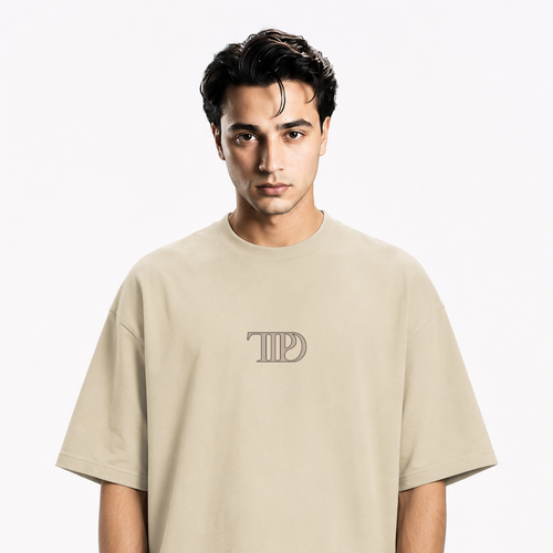 A man wearing an oversized beige crewneck t-shirt with a tonal 'TTPD' logo printed on the chest, set against a plain white background.
